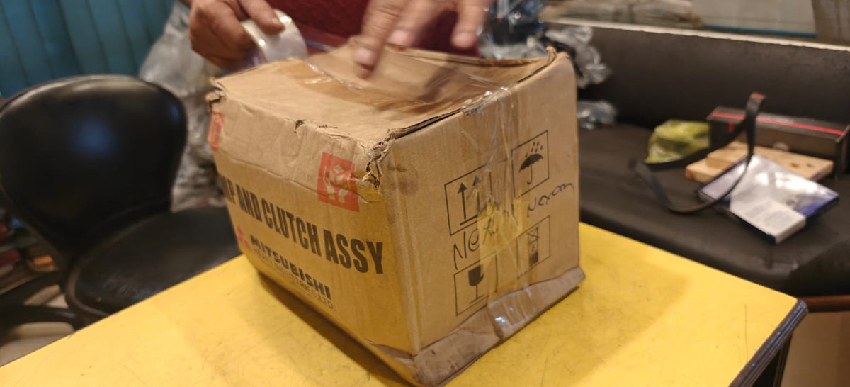 Cardboard box labeled 'Tata Xenon Air Compressor motor Mitsubishi' on a yellow surface with a blurred background.