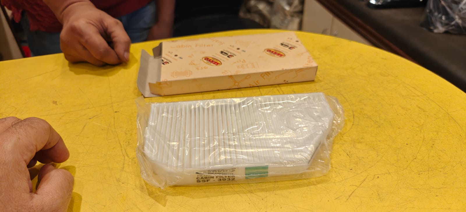 Packaged air filter on a yellow table with hands and a box in the background